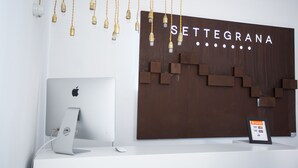 Un mostrador de recepción moderno con una iMac de Apple, una pared de madera con el letrero 'SETTEGRANA', lámparas colgantes y un expositor para tarjetas de pago.