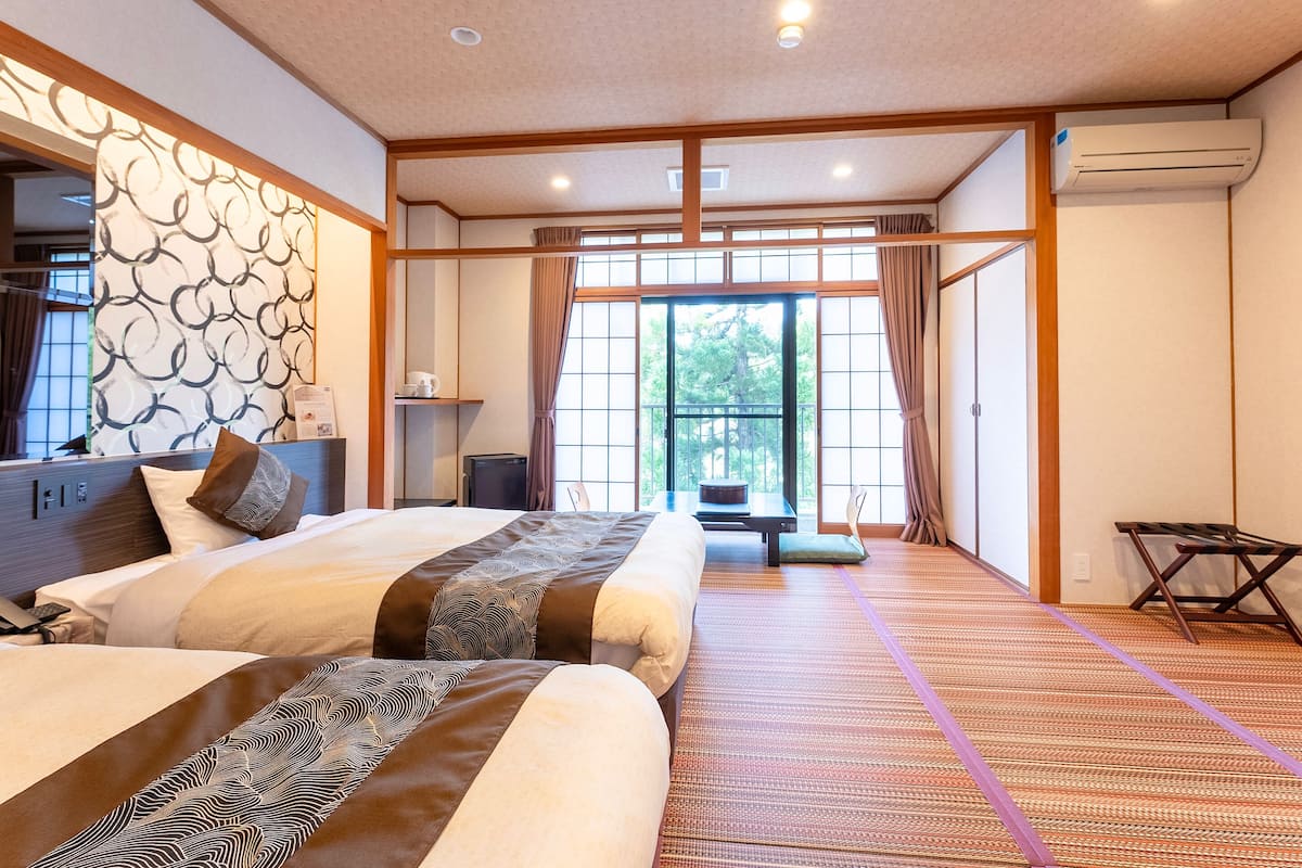 A traditional Japanese-style room with tatami flooring, a large window with a view of greenery, and a bed with patterned bedding.