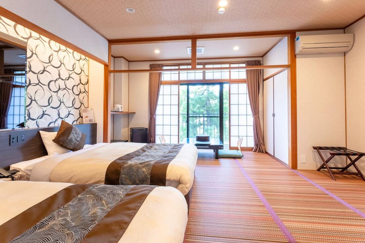A hotel room with two beds, a tatami mat floor, a sliding door, and a view of greenery outside.