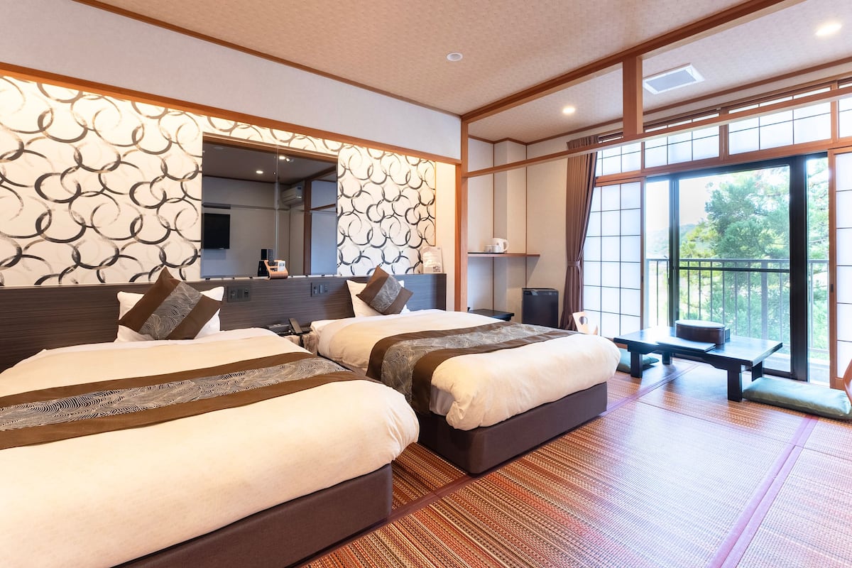 A hotel room with two beds, a large window, and a view of greenery.