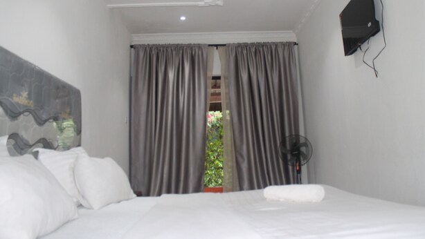 A hotel room with a bed, grey curtains, a wall-mounted TV, and a window with a view of greenery. - Honeybed Lodge (Lusaka)