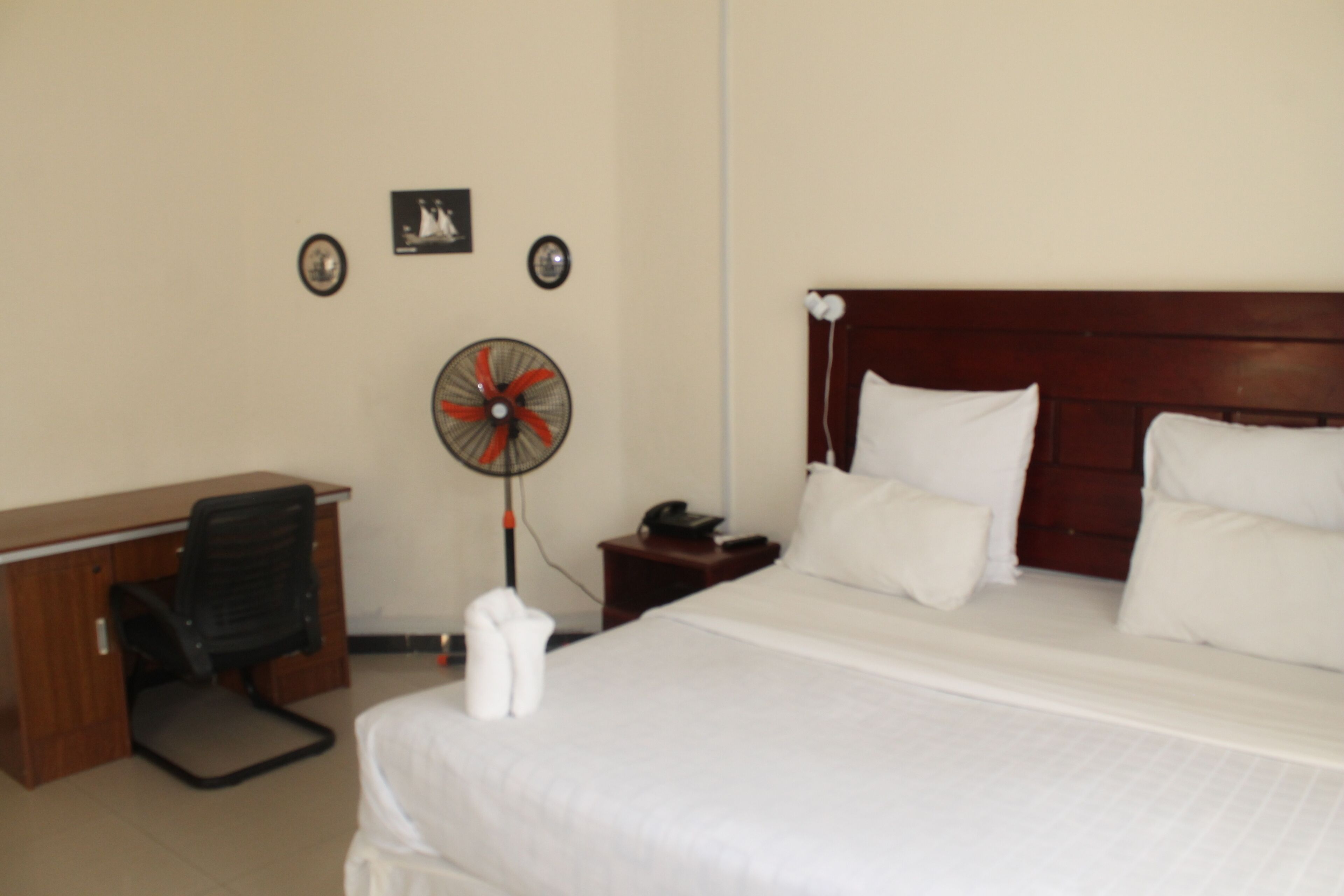 A hotel room with a bed, a desk, a chair, a fan, and a nightstand.