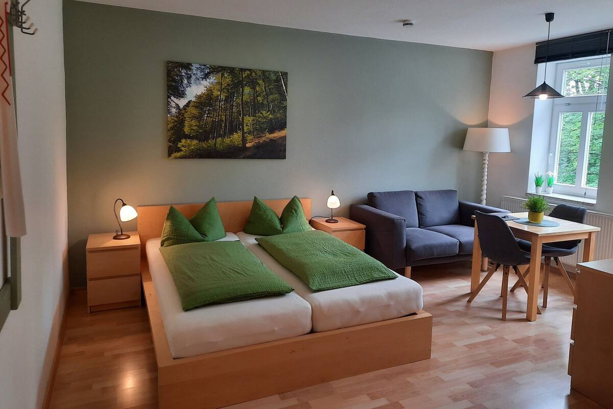 Une chambre moderne avec un lit en bois, des coussins verts, une table de chevet, une lampe de chevet, un canapé gris, une petite table avec une lampe et une fenêtre donnant sur un paysage verdoyant.