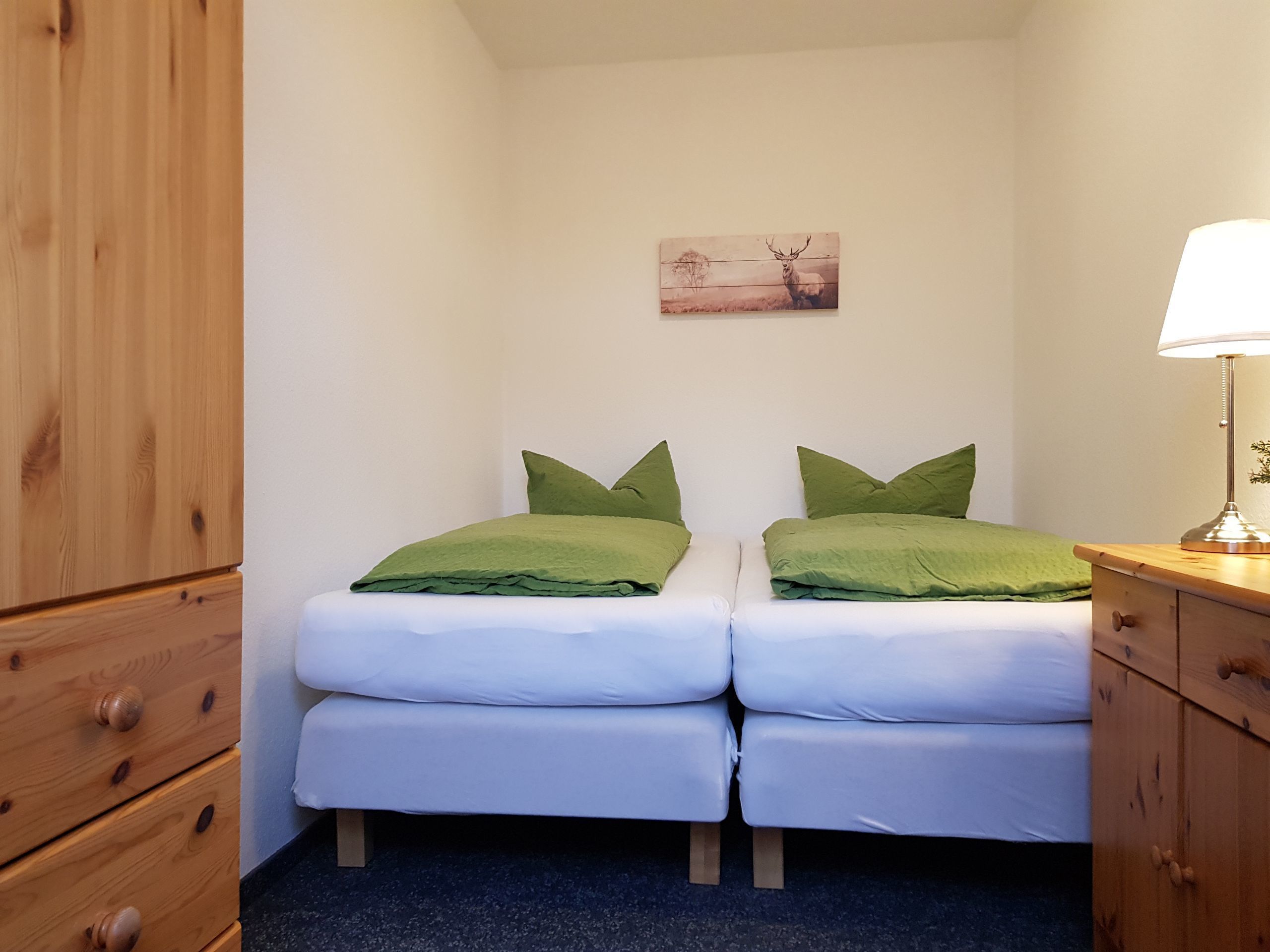 A bedroom with two single beds, a wooden wardrobe, a bedside table with a lamp, and a painting on the wall.
