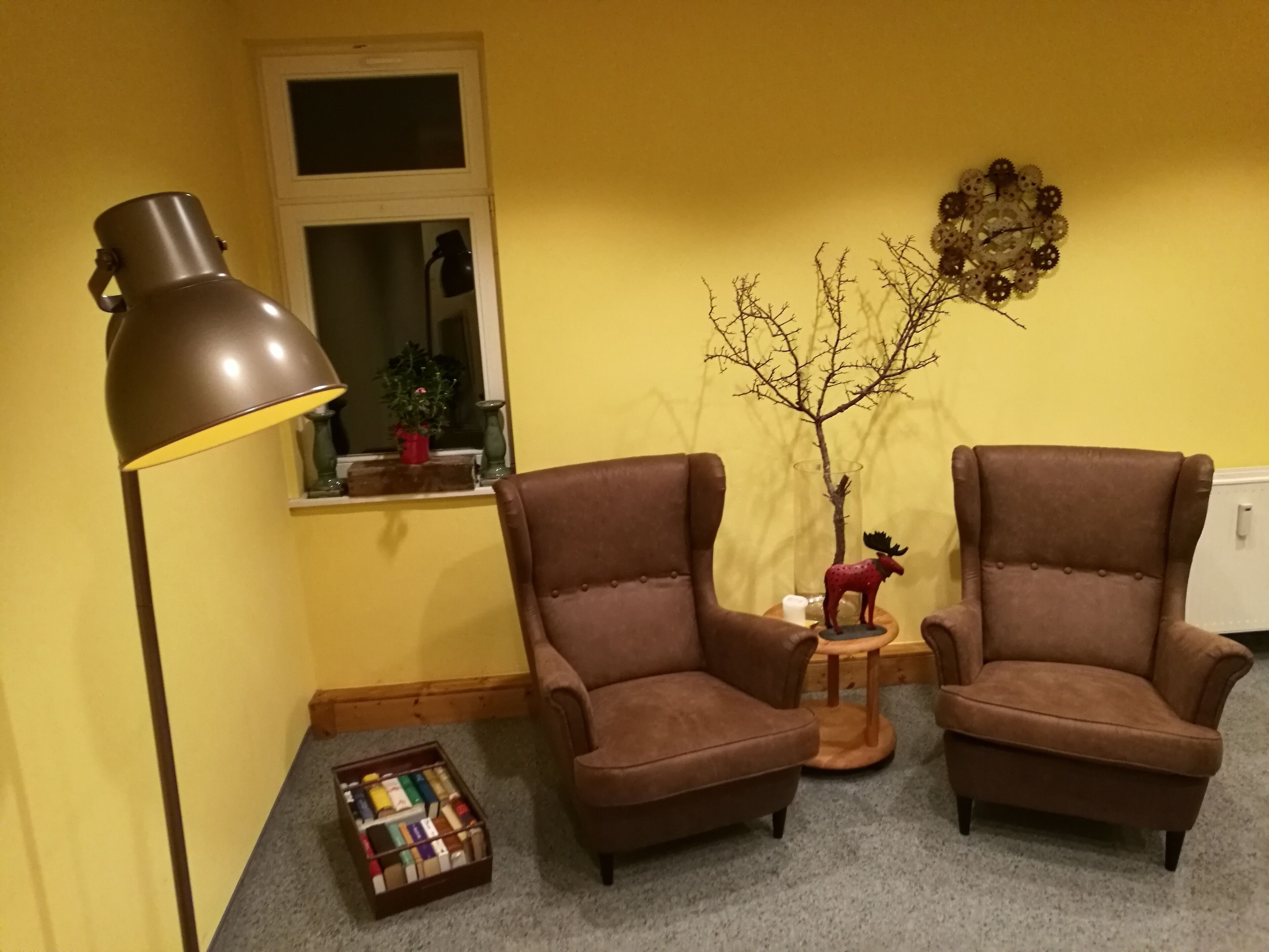 A room with two brown armchairs, a floor lamp, a small table with a moose figurine, a decorative tree, and a window with a view of the outside.
