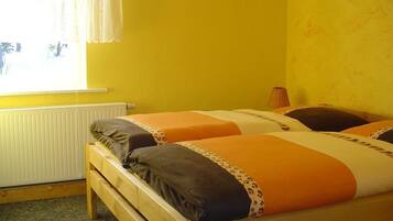 A room with two wooden beds, each with orange and white bedding, a radiator, and a window with white curtains.