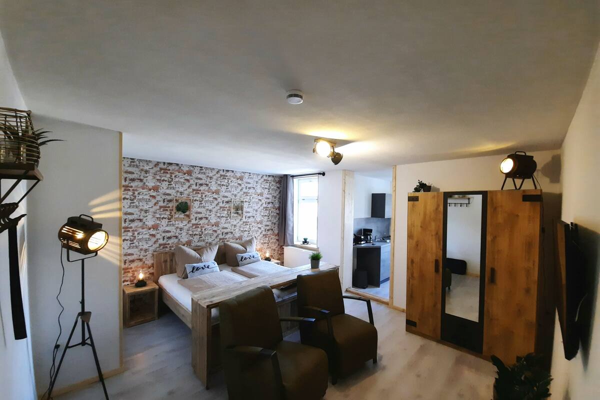 Une chambre à coucher de taille modeste, comprenant un lit, des tables de chevet, un bureau et une chaise. On aperçoit une petite kitchenette au fond.