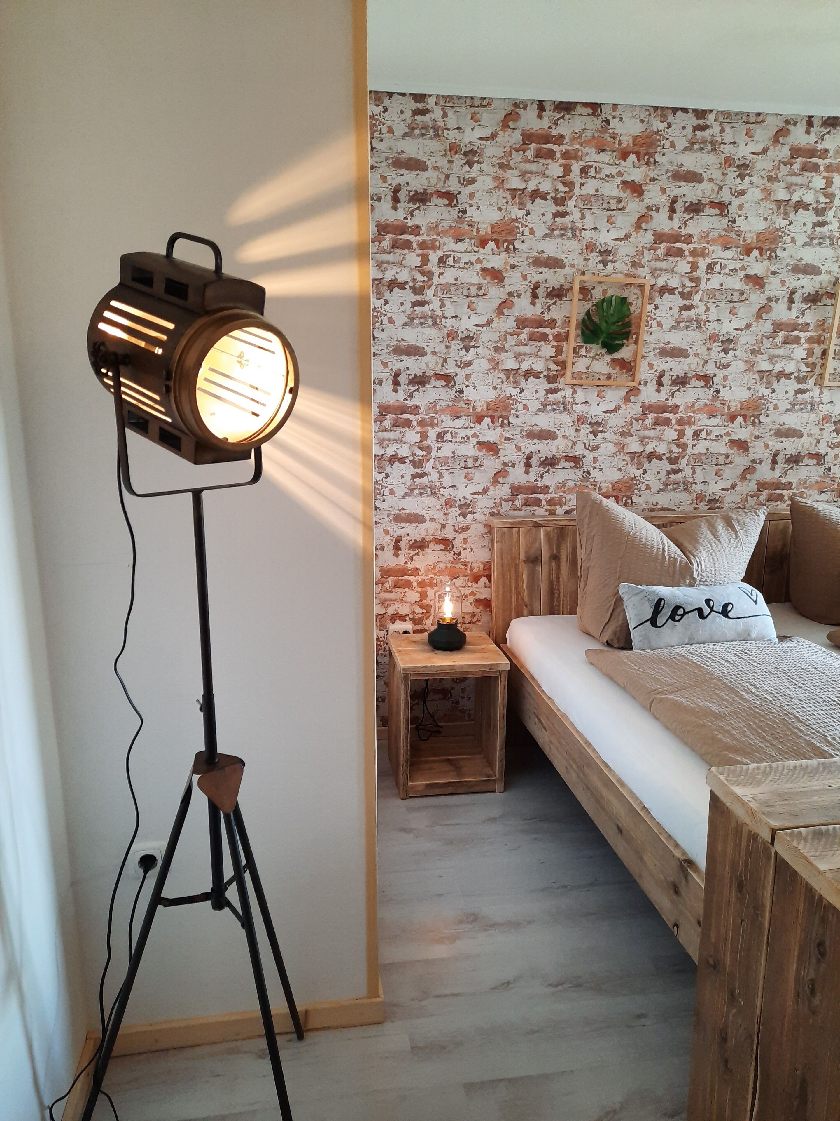 Un dormitorio con pared de ladrillo, una cama con un cojín que dice 'love', una mesita de noche con lámpara y una lámpara de pie.