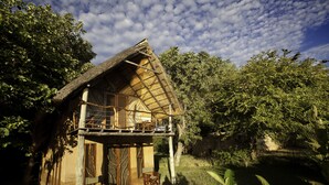 A rustic house with a thatched roof, a balcony, and a wooden chair outside. - Track and Trail River Camp (South Luangwa National Park)
