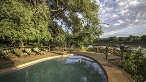 A swimming pool with lounge chairs and a wooden deck, surrounded by trees and a body of water. - Track and Trail River Camp (South Luangwa National Park)