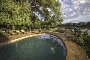 Piscina com espreguiçadeiras e deck de madeira, cercada por árvores e um corpo d'água.