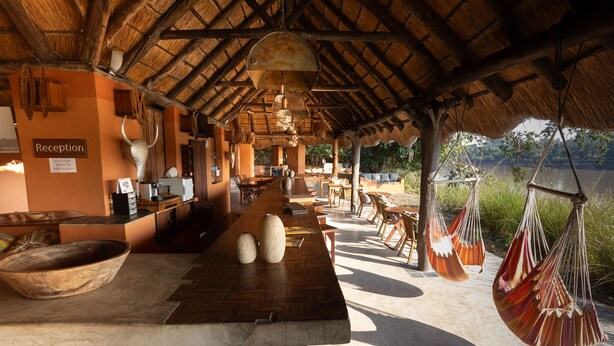 A reception area with a wooden reception desk, a stone wall, and a sign indicating no outside food allowed. There are hanging hammocks and a view of a body of water. - Track and Trail River Camp (South Luangwa National Park)