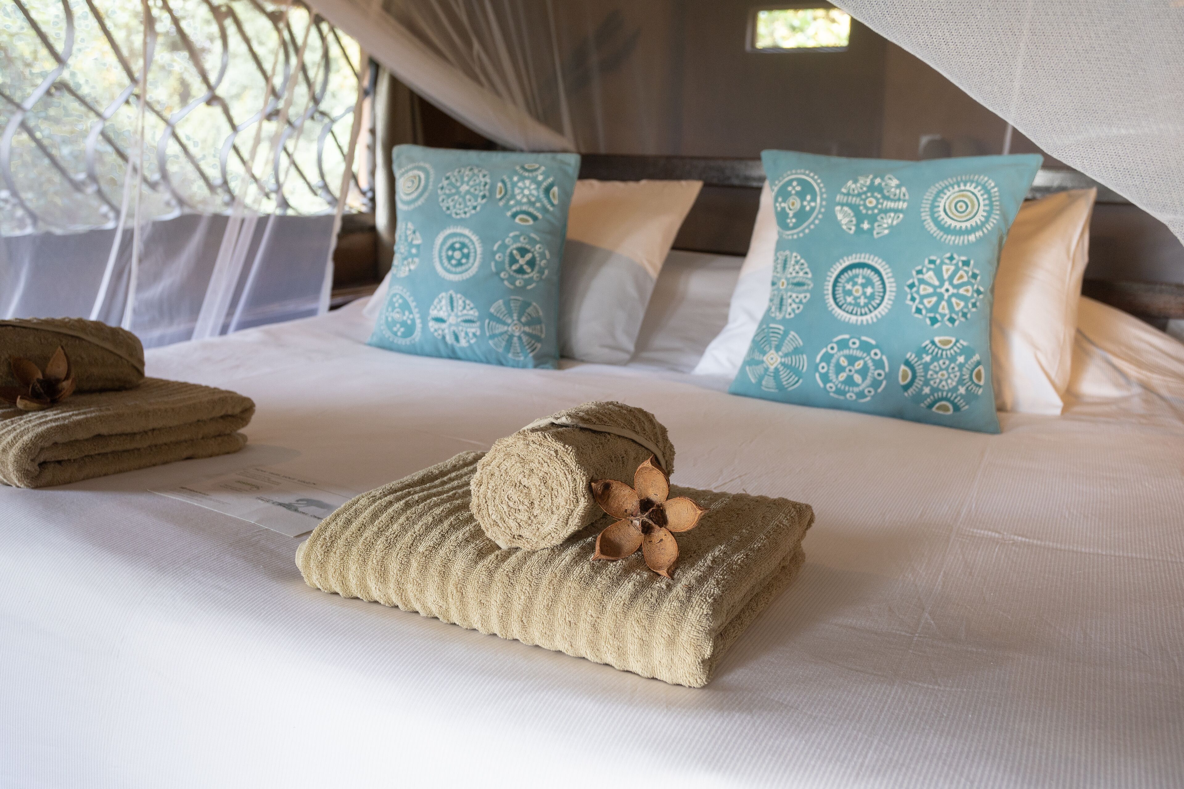 A neatly made bed with a rolled towel and decorative pillows, a window with a mesh curtain, and a wooden headboard.