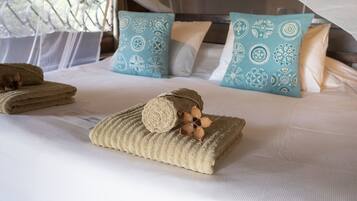 A neatly made bed with a rolled towel and decorative pillows, a window with a mesh curtain, and a wooden headboard.
