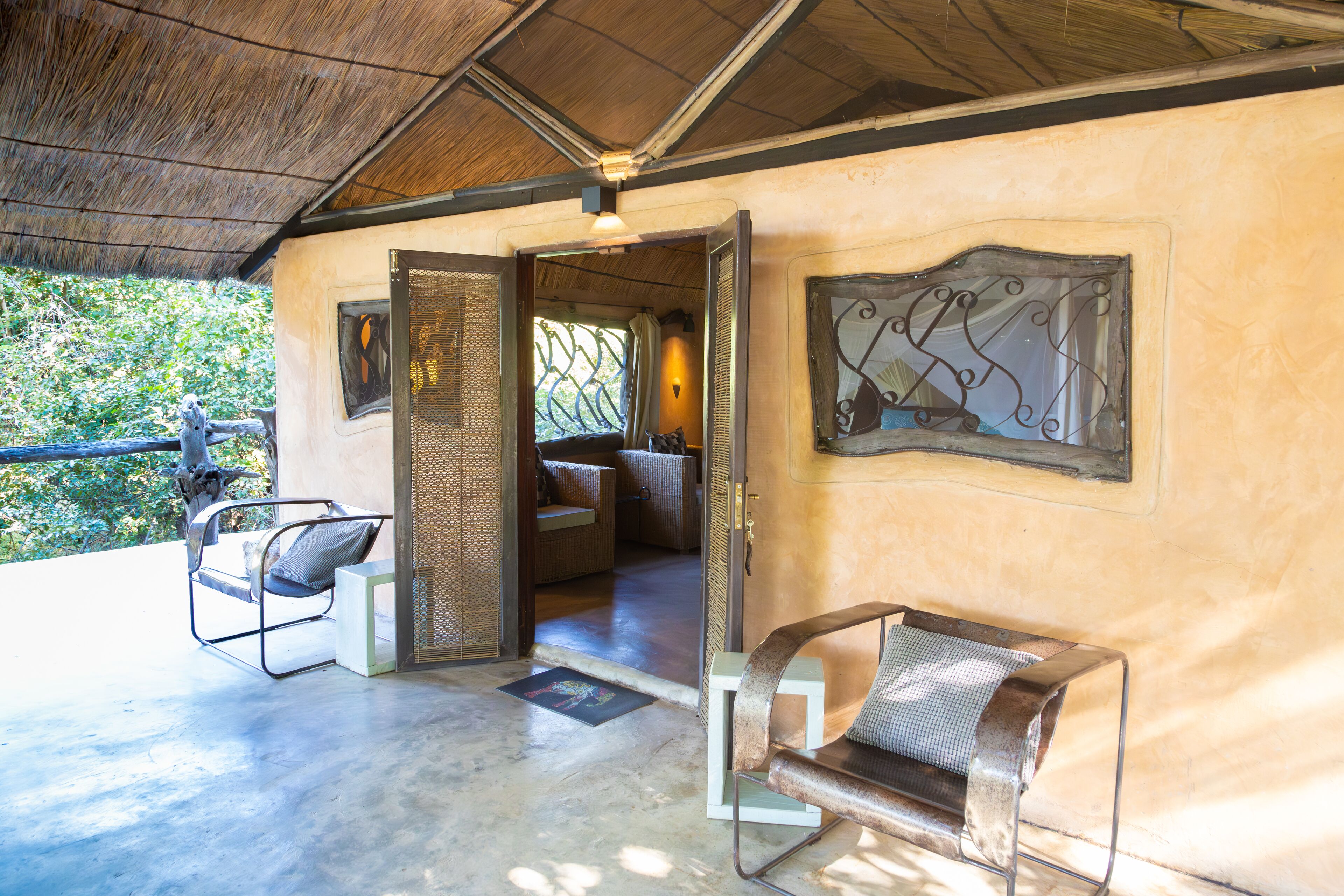 A covered outdoor seating area with metal chairs and a bench, a wooden door leading inside, and a decorative metal wall piece.