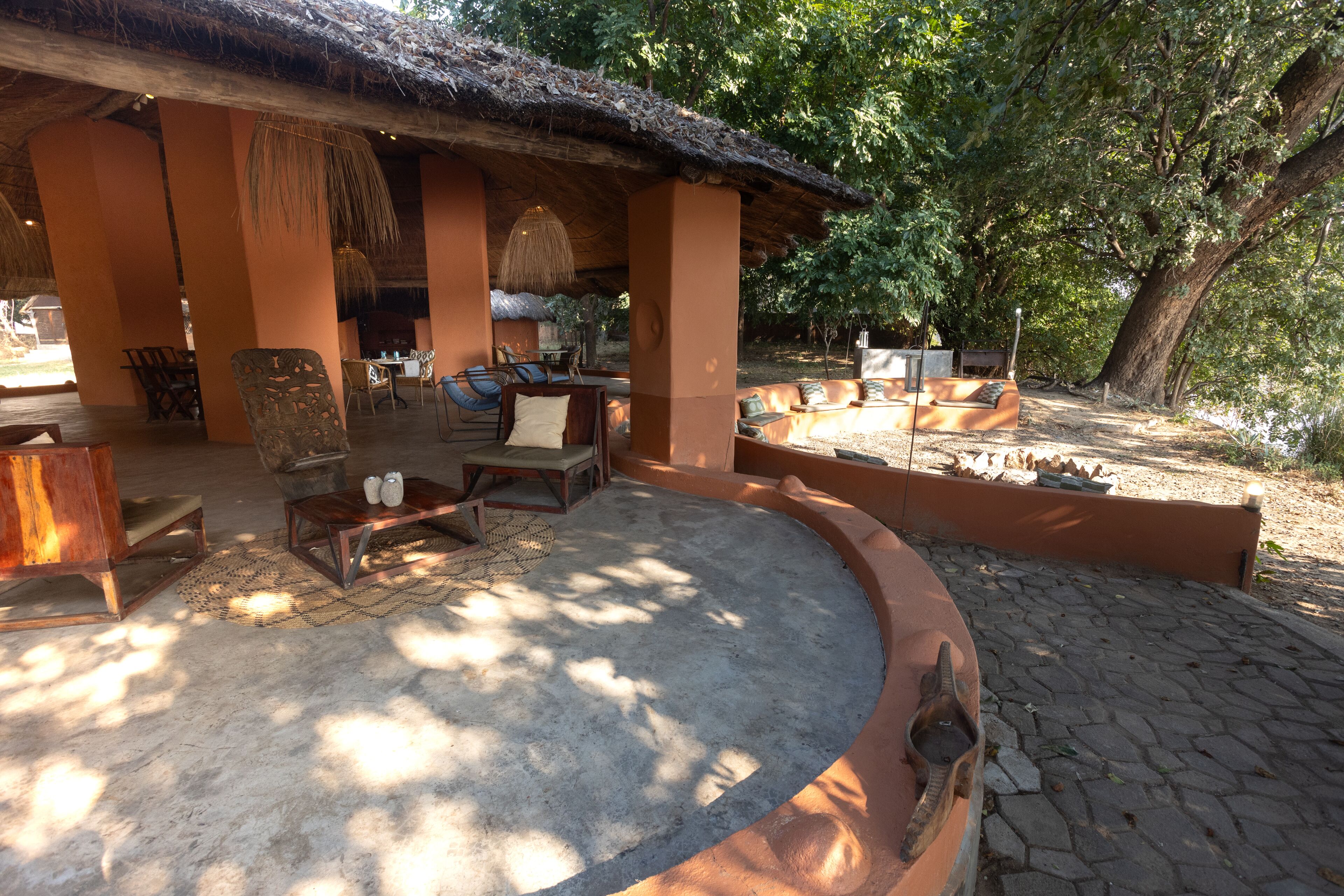 An outdoor seating area with a thatched roof, wooden furniture, and a stone-paved floor.