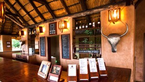 A rustic bar area with wooden flooring, a thatched roof, and a bull's head decoration. There are wine bottles on shelves and a menu board. - Track and Trail River Camp (South Luangwa National Park)
