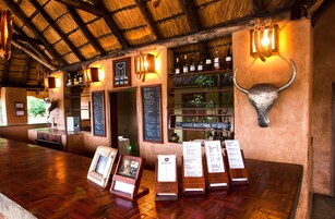 Uma área de bar rústica com piso de madeira, telhado de palha e decoração com cabeça de touro. Há garrafas de vinho em prateleiras e um quadro de menu.