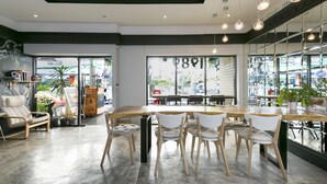 A modern cafe with a long wooden table, white cushioned chairs, and large windows offering a view of the street. - 1989 Sukhumvit Bangkok - Hostel (Bangkok)