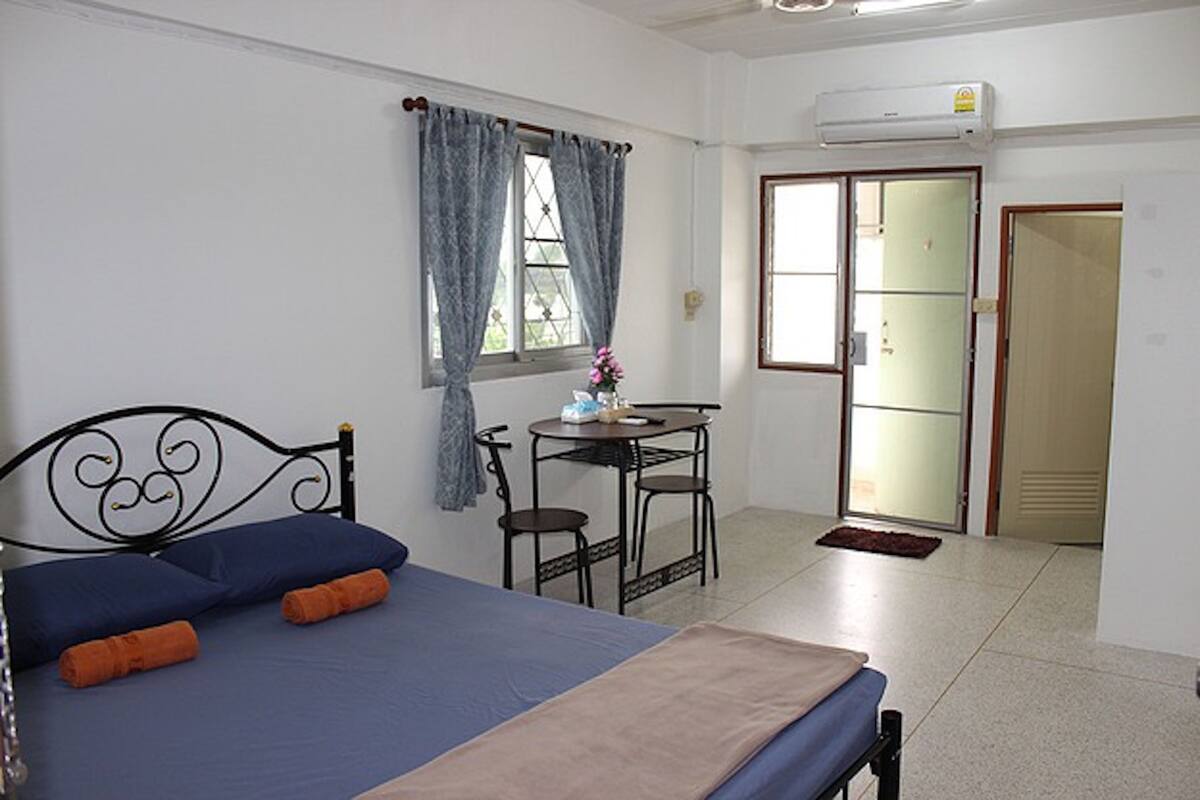 Un dormitorio con cama, escritorio y silla. Hay una ventana con cortinas, una puerta y un equipo de aire acondicionado en la pared.
