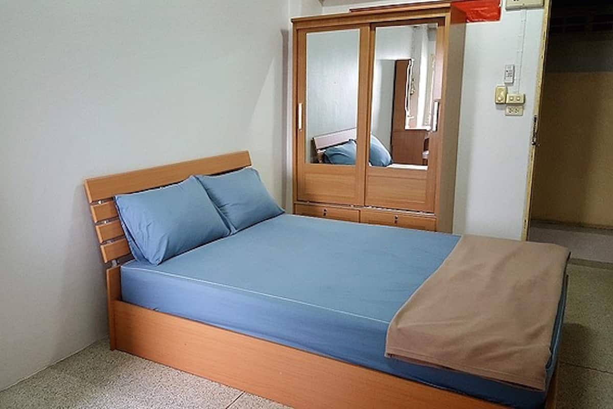 Una cama individual con cabecera de madera y ropa de cama azul, un armario de madera con espejo, y una manta color beige sobre la cama.