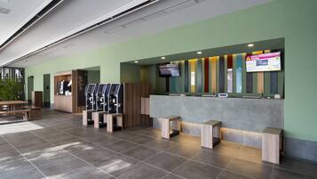 Een receptiezone met een groene wand, een digitaal scherm, een receptiebalie en een zithoek met houten krukken.