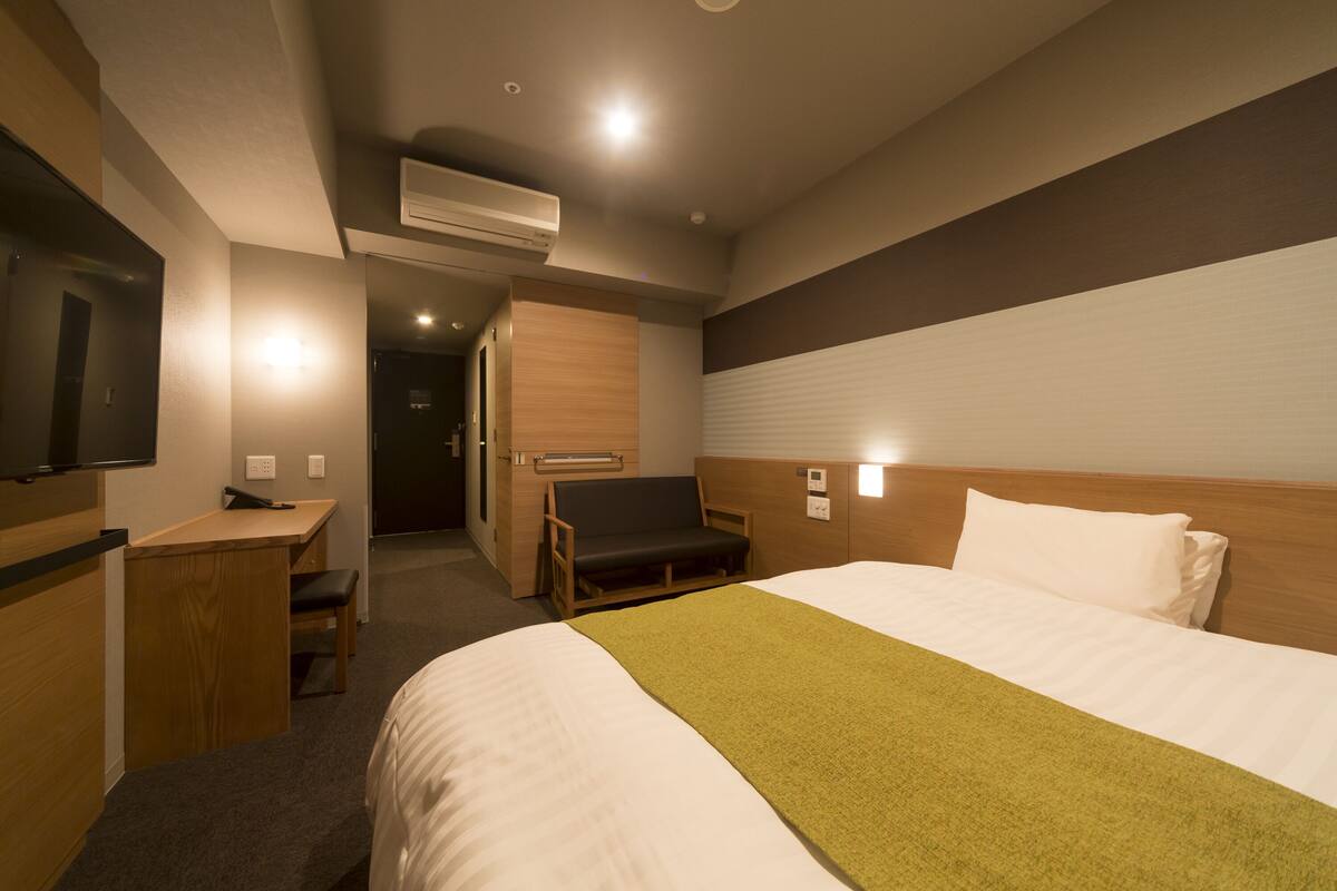 A hotel room with a bed, a desk, a chair, a TV, and an air conditioning unit.