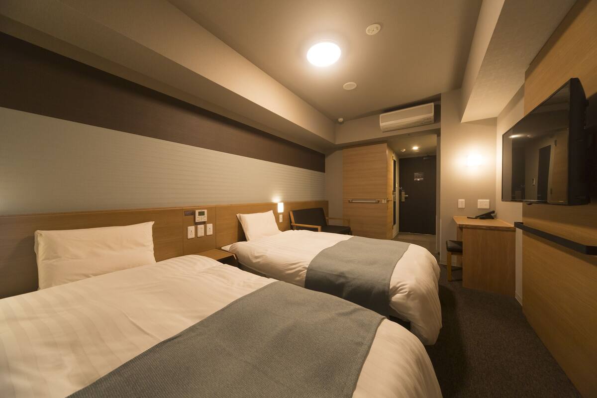 A hotel room with two beds, a desk, a chair, a TV, and an air conditioning unit.