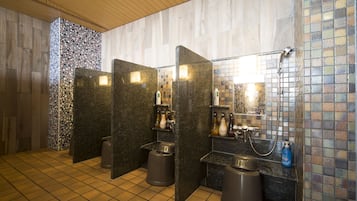 Baño público con tres cabinas, cada una con espejo y repisas para artículos de aseo. El suelo está cubierto de baldosas y hay botes de basura debajo de cada cabina.