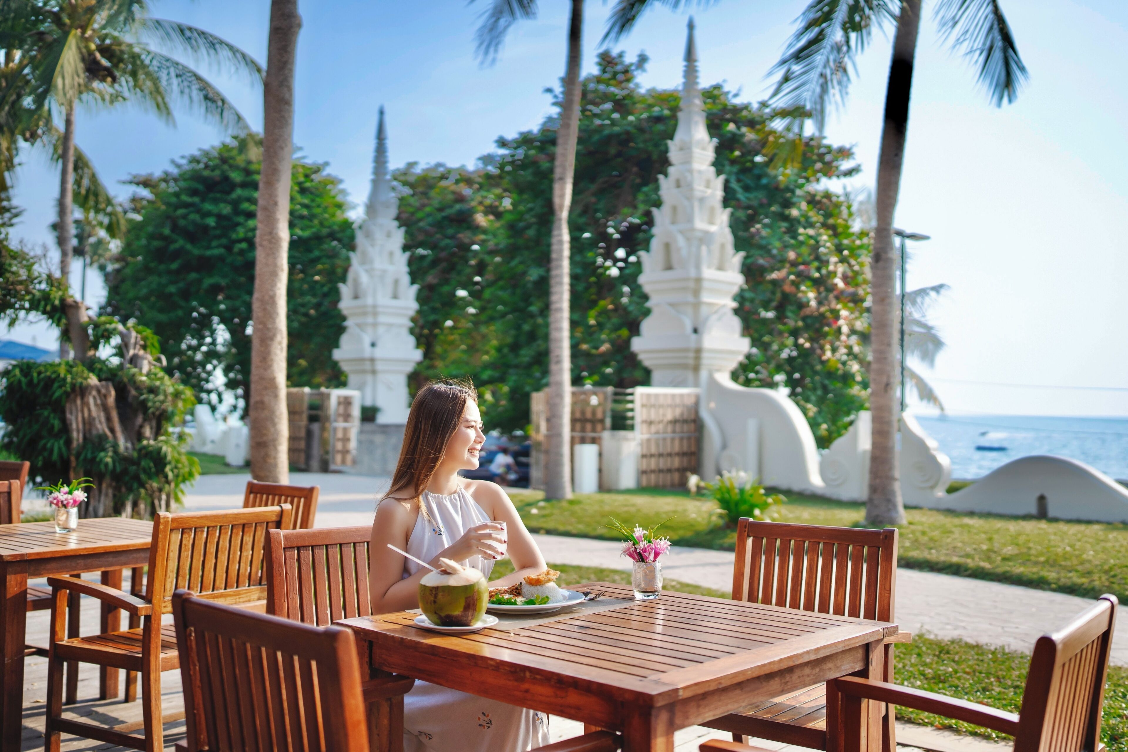 A woman dining at a wooden table with a view of white temple structures and the ocean.
