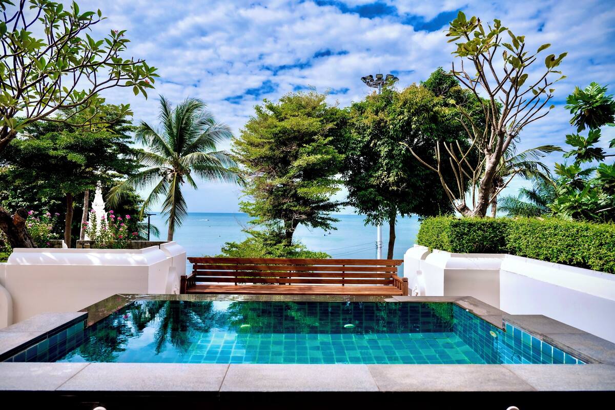 Une piscine à débordement avec une terrasse en bois, entourée d’une végétation luxuriante et offrant une vue sur l’océan.