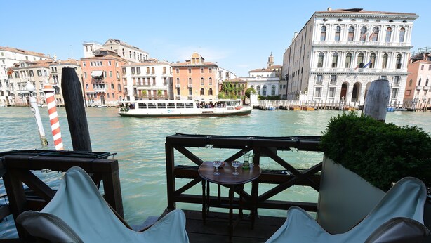 A riverside terrace with a table and chairs, overlooking a canal with historic buildings and a boat. - GranDELUX-Canal (Venice)