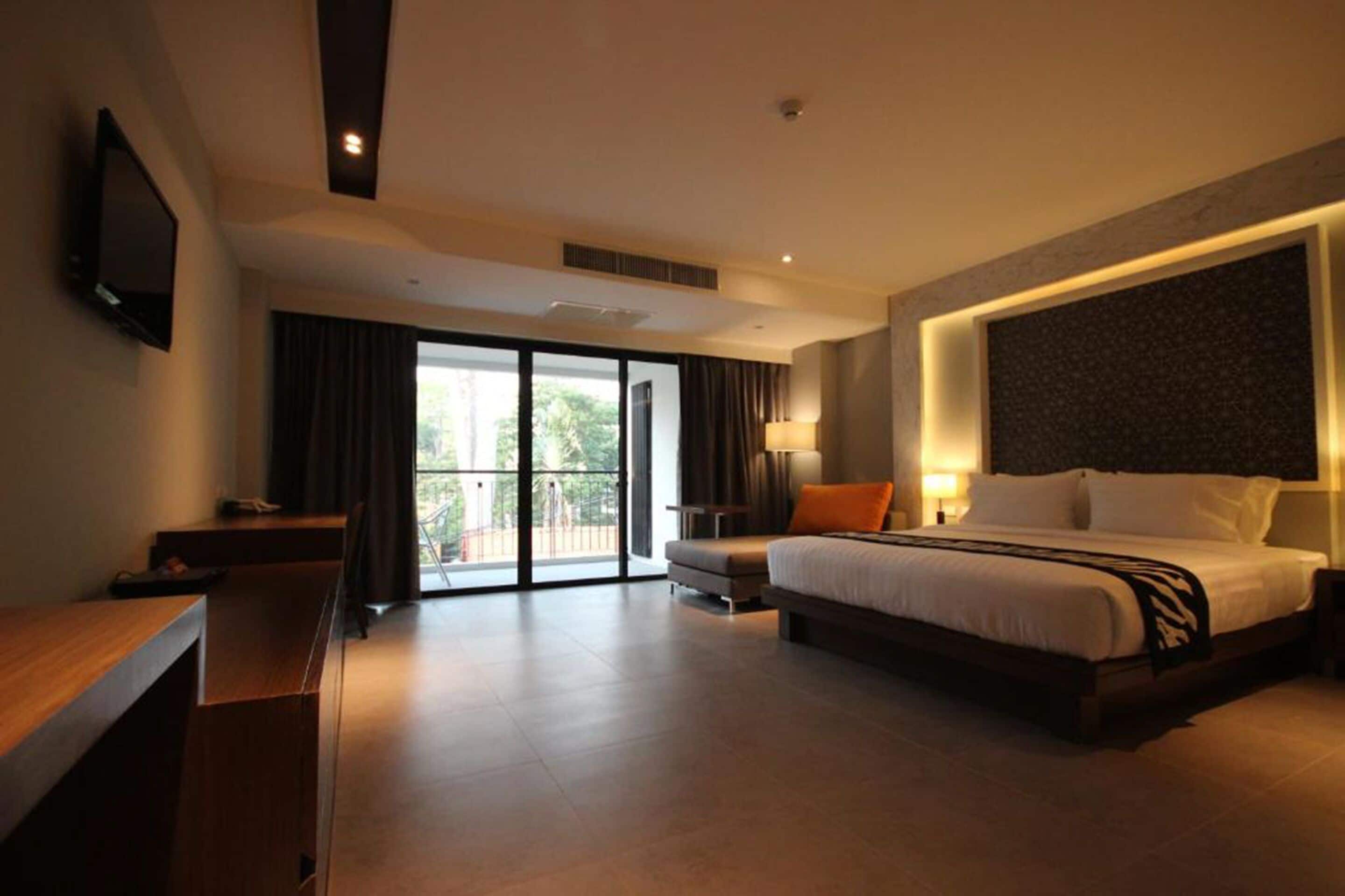 A modern hotel room with a large bed, a desk, a TV, and a balcony with curtains.