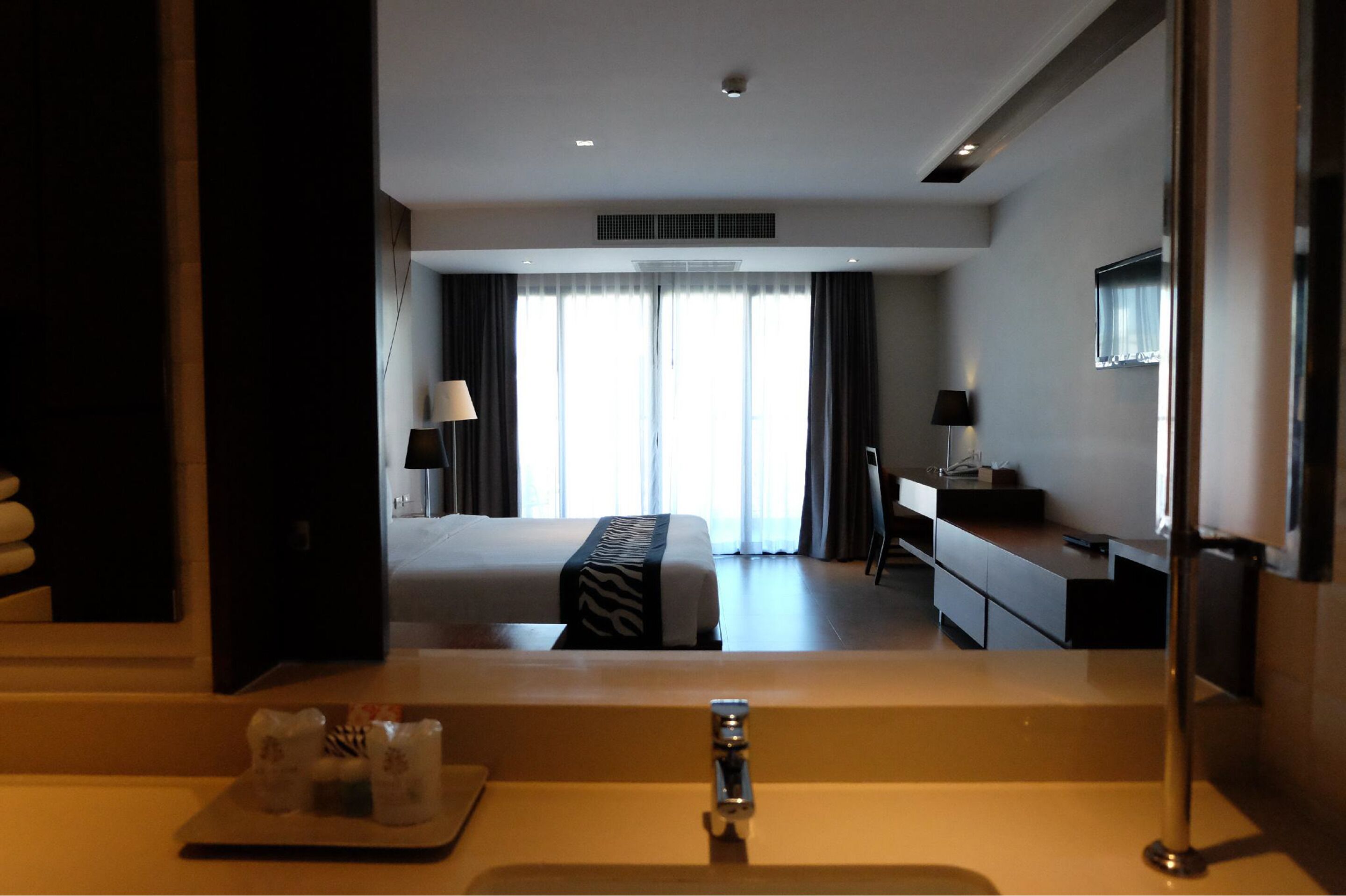 A hotel room with a bed, a desk, a TV, and a bathroom with a sink and mirror.