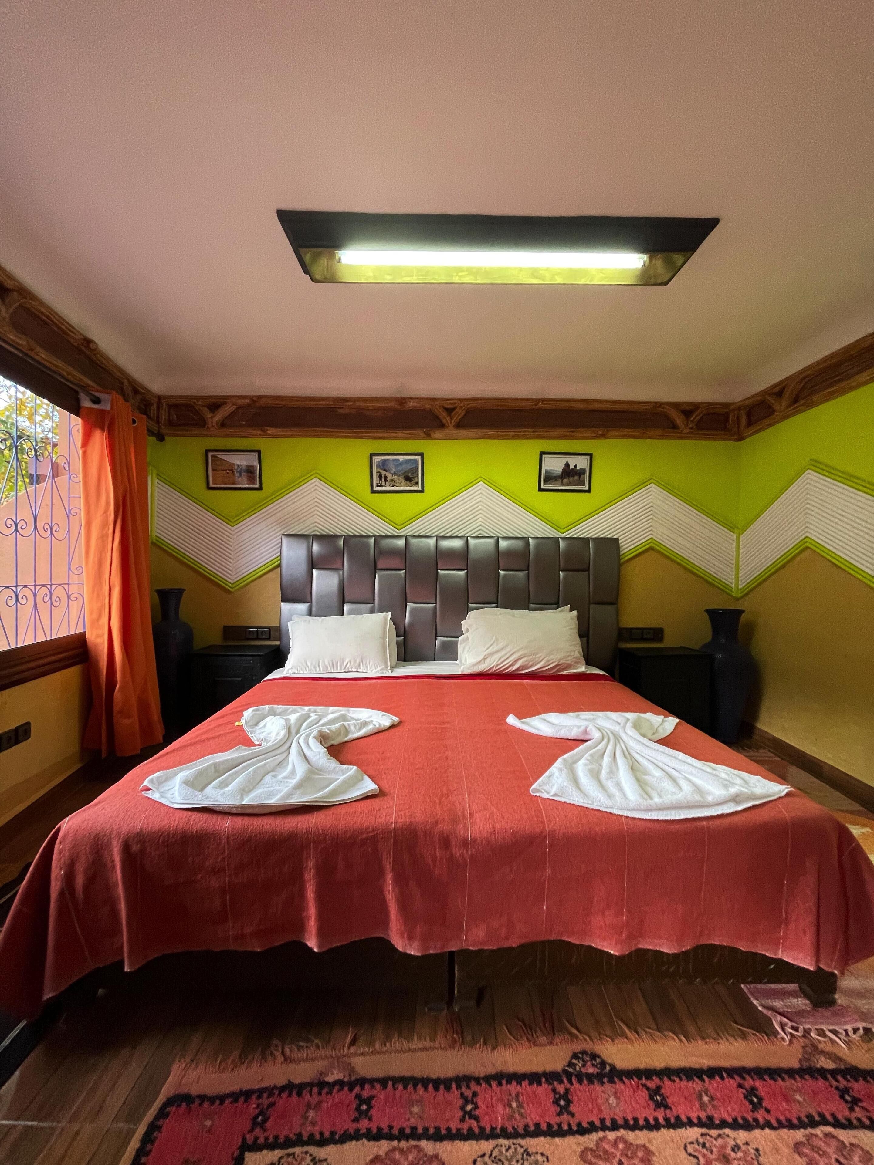 A hotel room with a bed, two pillows, a headboard, a nightstand, a vase, a picture frame, a window with curtains, and a patterned rug.