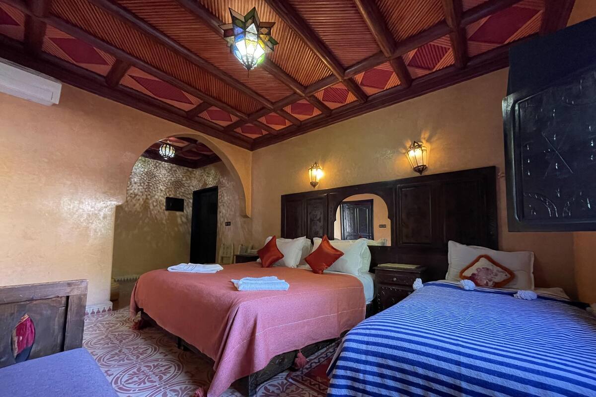 Quarto de hotel com duas camas, piso estampado, vigas de madeira no teto e um lustre decorativo.