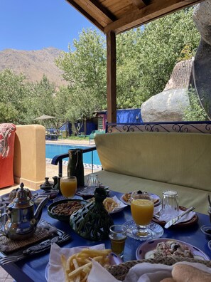 A dining table set with various dishes and drinks, overlooking a pool and mountains.