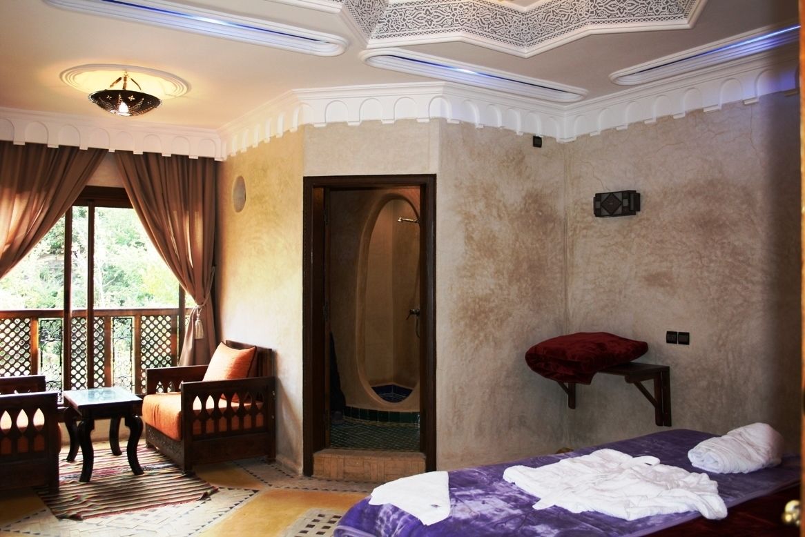 Una habitación con cama, una mesita, dos sillas, un ventanal con cortinas y una puerta que da a un baño.
