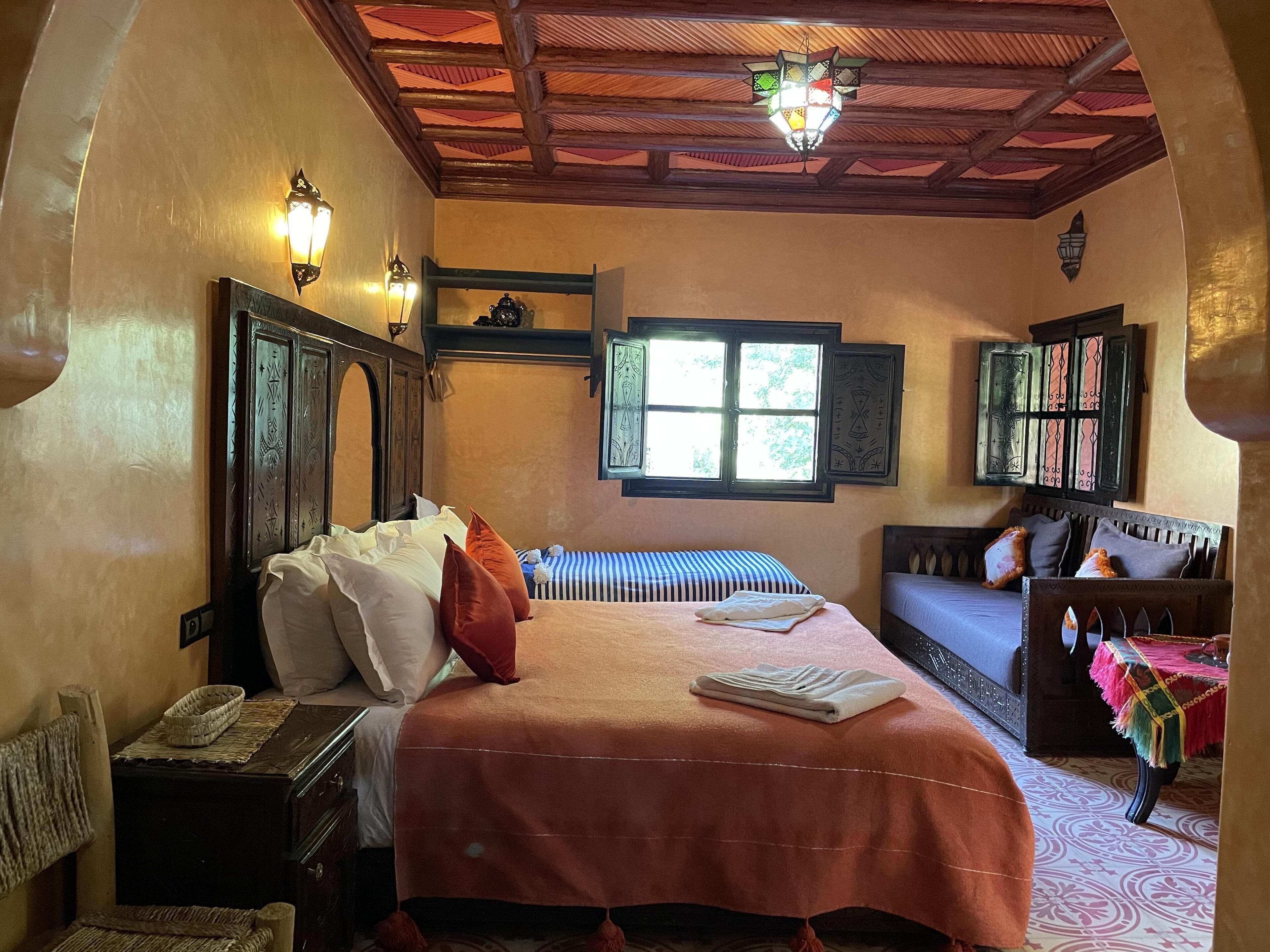 Una habitació acollidora amb un llit gran, un panell de capçalera de fusta, una finestra amb persianes decoratives i una taula petita amb un mantell de taula de colors.