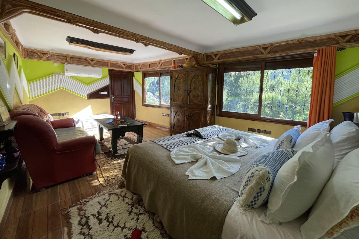 Quarto com cama, poltrona vermelha, armário de madeira e janela com vista para o verde.