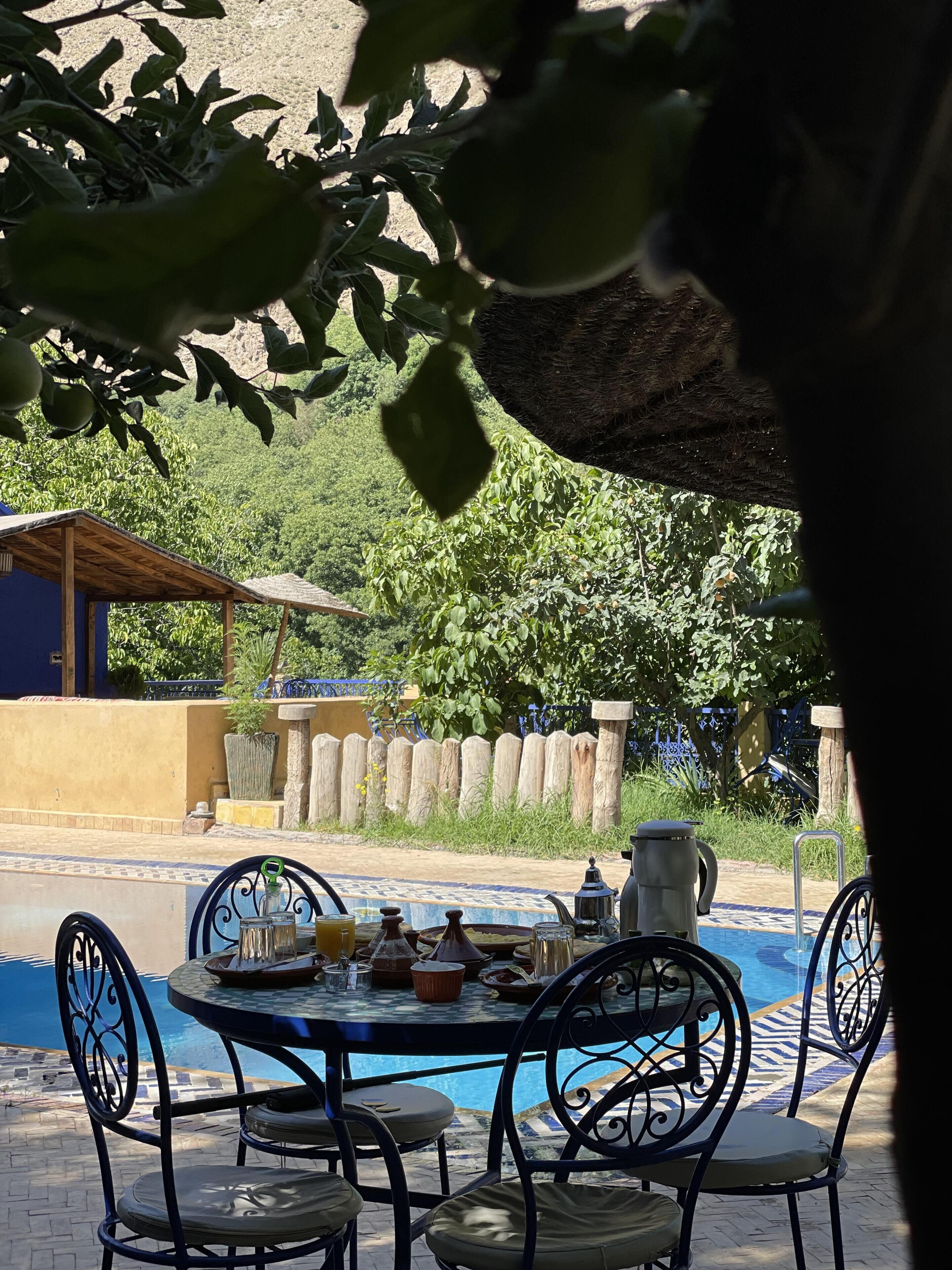 Zona de piscina con mesa lista para comer, rodeada de sillas y vista a una piscina y follaje al fondo.