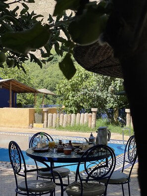 A poolside area with a table set for dining, surrounded by chairs, and a view of a pool and greenery in the background.