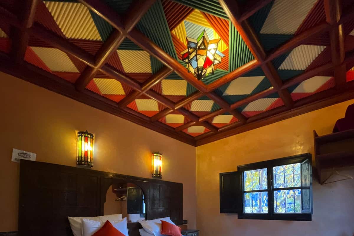 Quarto de hotel com uma cama grande, duas mesas de cabeceira, um criado-mudo, uma janela com vista e teto decorado.