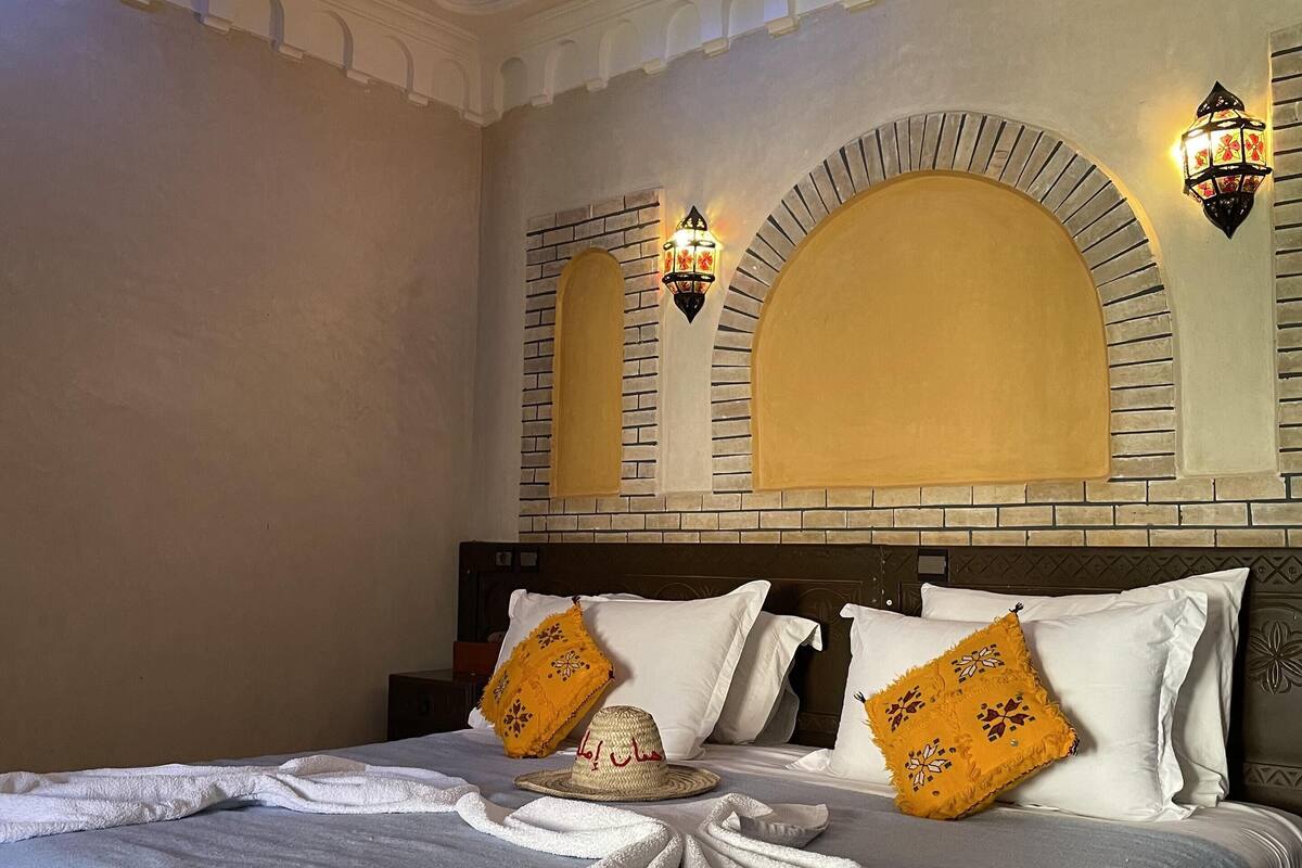 Quarto de hotel com uma cama grande, almofadas decorativas, cabeceira com desenho estampado e luminárias fixadas na parede.