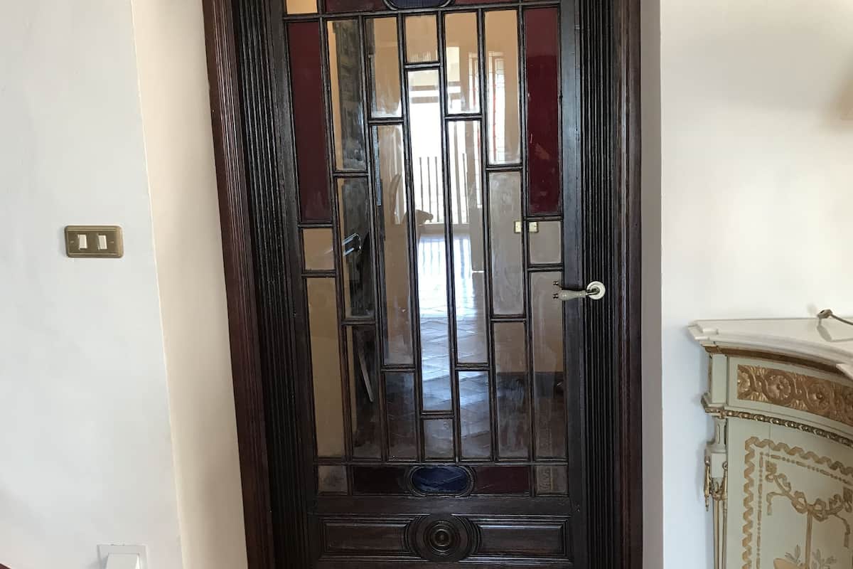 Uma porta de madeira escura com painéis de vidro e um detalhe decorativo no topo, dando acesso a um chão de azulejos com borda estampada.