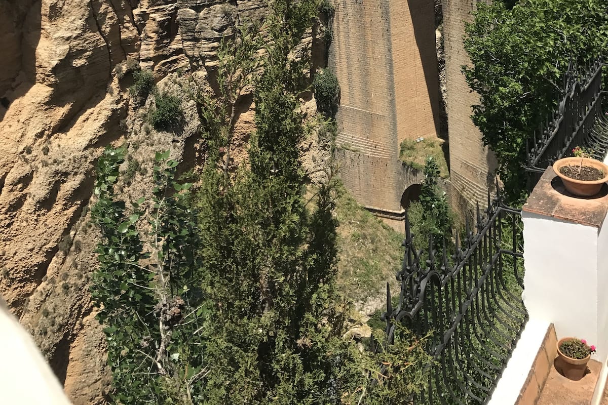 Uma ponte de pedra histórica com arcos, terreno rochoso com vegetação escassa e uma fileira de edifícios ao fundo.