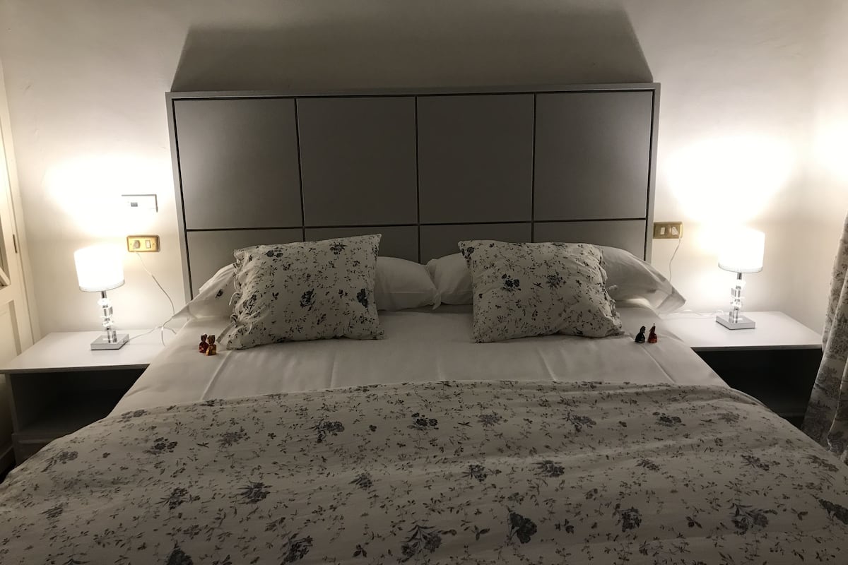 Cama bem arrumada com roupa de cama florida, duas luminárias de cabeceira e cabeceira com padrão quadriculado.