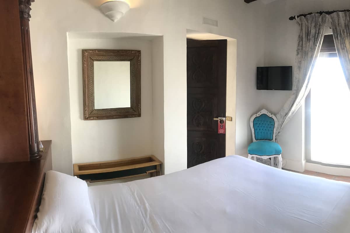 Quarto com cama, espelho, televisão, cadeira e janela com cortinas.