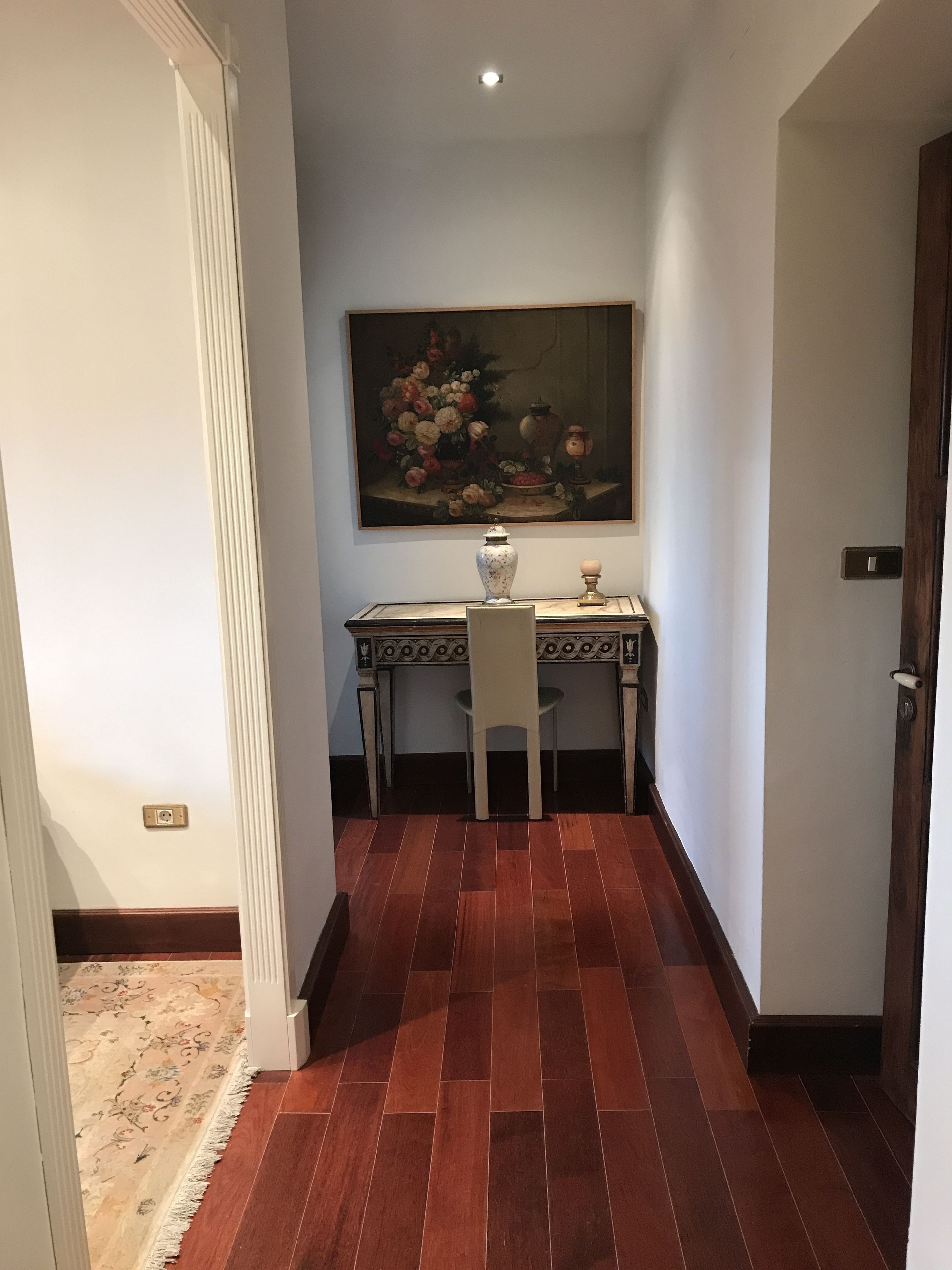 A hallway with a dining table and chairs, a vase, and a painting on the wall.