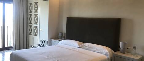 A modern hotel room with a large bed, a nightstand, a lamp, and a view of a balcony with curtains.
